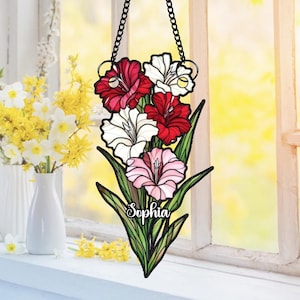 Könnte beinhalten: Ein Fensterbehang aus Glas mit roten, weißen und rosa Gladiolenblüten und grünen Blättern. Der Name "Sophia" ist auf den Blättern geschrieben. Aufgehängt an einer schwarzen Kette.