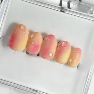 May include: Five artificial nails with a peach and pink gradient design, each featuring a clear, droplet-shaped detail. The nails are presented in a clear plastic case.