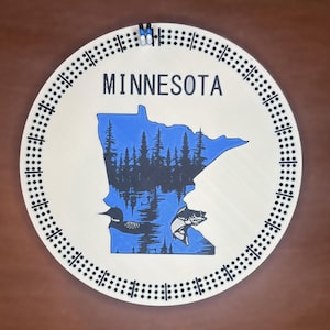 May include: A round wooden cribbage board featuring the state of Minnesota in blue, with a black forest and lake scene. The word "MINNESOTA" is printed above the state outline. The board has a track of drilled holes for pegs.