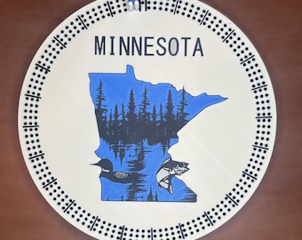 May include: A circular wooden cribbage board with the state of Minnesota in blue, and a black forest and lake scene. The word "MINNESOTA" is printed above the state outline. The board has a track of drilled holes for pegs.