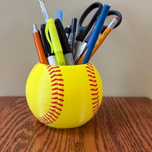 May include: A yellow softball-shaped desk organizer filled with pens, pencils, and scissors. The organizer has red stitching details and sits on a wooden surface. The container holds various writing utensils.
