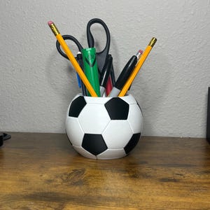 Può includere: Un organizer da scrivania a forma di pallone da calcio bianco e nero, pieno di matite gialle, forbici nere e varie penne e pennarelli. L'organizer è posto su una superficie di legno.