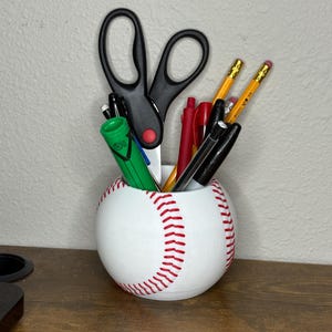 May include: A white baseball-shaped desk organizer with red stitching. The organizer holds black scissors, pens, pencils, and a green marker. The desk accessory sits on a brown wooden surface.