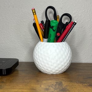 Op de afbeelding: Een witte bureau-organizer in de vorm van een golfbal, gevuld met pennen, potloden en scharen. De organizer is gemaakt van een textuurrijk materiaal en staat op een houten oppervlak. Een groene markeerstift is zichtbaar.