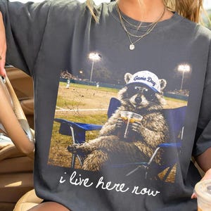 May include: Dark gray t-shirt featuring a raccoon wearing sunglasses and a "Game Day" hat, seated in a blue chair, holding a drink, with a baseball field backdrop. The text "i live here now" is printed below the image.