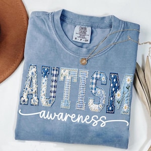May include: A light blue Comfort Colors t-shirt with the word "AUTISM" in patchwork letters, featuring floral and gingham patterns. The word "awareness" is written below in white script. A gold necklace with a pendant rests on the shirt.