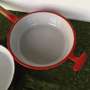 May include: A red enamel cast iron skillet with a wooden handle and a white interior. The skillet is sitting on a green patterned surface.