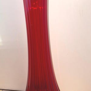 May include: A tall, red glass vase with a flared, ruffled top and a ribbed design. The vase is sitting on a wooden surface.