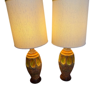 May include: Pair of vintage table lamps. Each lamp features a beige fabric shade with a green trim. The bases are brown with yellow and green accents. The lamps are on a dark brown wooden base.