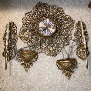 May include: A gold-toned wall clock with ornate metal accents and two matching candle sconces. The clock has a round face with Roman numerals and a chain hanging from the bottom.