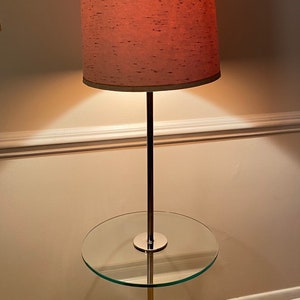 May include: A chrome floor lamp with a pink fabric shade and a glass side table. The lamp has a round base and a long, slender stem.