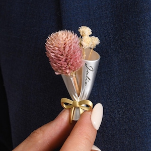 May include: A small bouquet brooch featuring dried flowers in a silver cone, tied with a gold ribbon. The cone has the name "Julia" written on it. The brooch is held against a navy blue blazer.