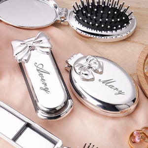 Puede incluir: Espejo compacto plateado, peine y dos estuches con lazos decorativos. Un estuche rectangular con "Avery", el otro ovalado con "Mary". También se ve un pequeño anillo dorado con una piedra rosa.