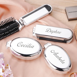 Op de afbeelding: Een verzameling zilverkleurige compacte spiegels en een opvouwbare haarborstel met spiegel. Drie ovale compacts zijn gegraveerd met de namen "Cecilia", "Arabela" en "Daphne". De haarborstel heeft een zwart borstelkussen. Er is ook een kleine vierkante doos aanwezig.