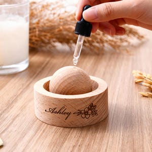 May include: A wooden essential oil diffuser with a wooden sphere inside a round holder. The holder is engraved with the name "Ashley" and a floral design. A dropper is held above the sphere, dispensing liquid. A candle and dried plants are in the background.
