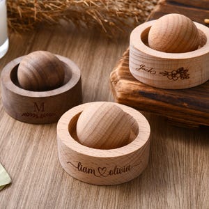 May include: Three wooden ring holders with a ball inside. One is dark brown with the letter "M" and a floral design. Another is light brown with the names "Liam & Olivia" and a heart. The third is light brown with the name "Jack" and a floral design.