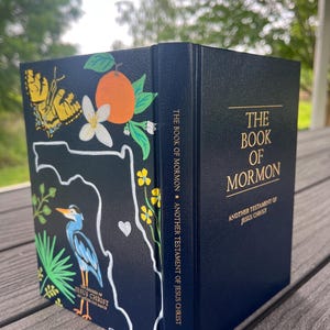 May include: A navy blue book titled "The Book of Mormon" with gold lettering. The cover features a hand-painted design with a butterfly, orange, flowers, and a blue heron. The book's spine reads "The Book of Mormon - Another Testament of Jesus Christ."