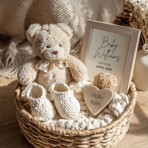 Puede incluir: Un oso de peluche beige, patucos de bebé y un corazón de madera con "Hello Baby" están en una cesta de mimbre. Una impresión enmarcada dice "Baby Williams ARRIVING APRIL 2026". La escena está sobre una manta de punto.