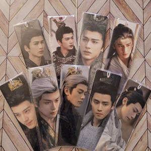May include: A collection of nine rectangular bookmarks, each encased in a clear plastic sleeve, featuring photographic portraits of a person in various traditional East Asian-inspired costumes, hairstyles, and headpieces, with white tassels attached.