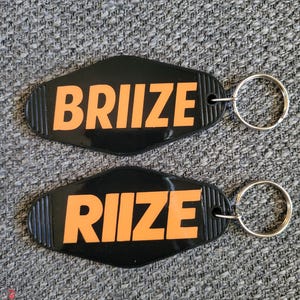 May include: Two black keychains with orange text that reads "BRIIZE" and "RIIZE". The keychains have a retro motel-style design with a silver key ring attached. The background is a textured gray surface.