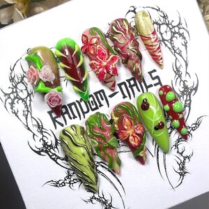 Puede incluir: Un conjunto de diez uñas artificiales largas en forma de almendra. Cada uña presenta un diseño único, incluyendo patrones florales, purpurina y arte abstracto en tonos verdes, rojos, rosas y dorados. El texto "RANDOM-NAILS" es visible.