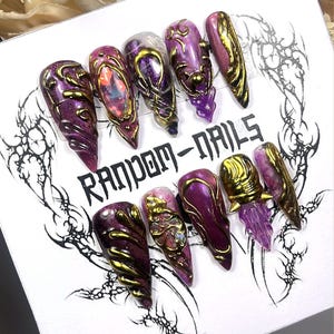 Puede incluir: Un conjunto de diez uñas artificiales largas y puntiagudas. Cada uña es de color púrpura intenso con detalles dorados y diseños ornamentados. El texto "RANDOM-NAILS" está impreso en la superficie blanca.