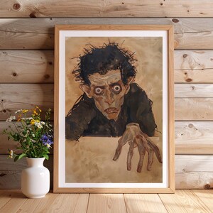 May include: Framed artwork featuring a watercolor painting of a gaunt figure with dark hair and a somber expression. The figure's hand is outstretched. The artwork is in a light wood frame, set against a wooden background.