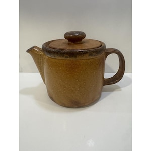 May include: A cylindrical ceramic teapot with a speckled brown glaze, featuring a matching lid with a central knob, a curved handle on the right side, and a short, angled spout on the left. The surface has a textured, earthy appearance.