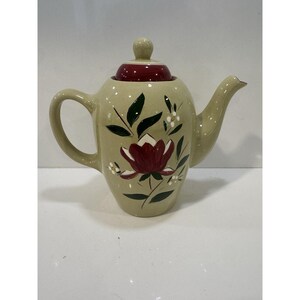 May include: A beige ceramic teapot with a burgundy lid and a floral design. The teapot features a large burgundy flower with green leaves and small white flowers. The handle and spout are curved.