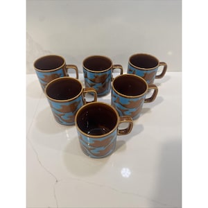 May include: Six ceramic coffee mugs. Each mug has a brown interior and handle, with a light blue exterior featuring a brown fish design. The mugs are arranged in a circular pattern.