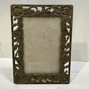 May include: Ornate bronze-colored picture frame with floral and scrollwork detailing. The rectangular frame features a central opening for a photograph or artwork. The corners and edges are embellished with sculpted flowers and leaves.