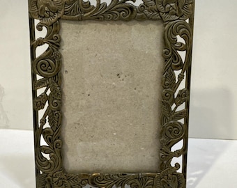 May include: Ornate bronze-colored picture frame with floral and scrollwork detailing. The rectangular frame features a central opening for a photograph or artwork. The corners and edges are embellished with sculpted flowers and leaves.