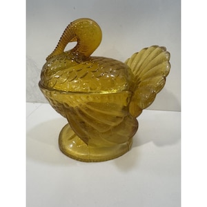 May include: Amber-colored glass turkey-shaped candy dish with a lid. The dish features detailed feather and body patterns, a curved neck, and a fan-shaped tail. The base is round and the dish is elevated.