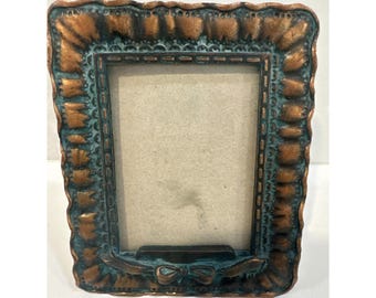 May include: Ornate bronze-colored picture frame with a rectangular opening. The frame has a distressed finish with teal accents and a decorative bow at the bottom. The frame's edges are scalloped.