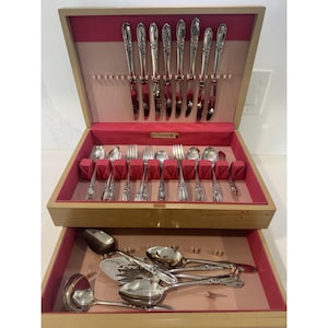 May include: A wooden silverware chest with a pink interior, holding a complete set of silver utensils. The top section displays knives, forks, and spoons, while the bottom drawer contains serving pieces. The silverware has ornate handles.