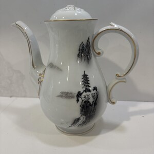 May include: White porcelain coffee pot with a lid, featuring a black and gray landscape design. The pot has a curved handle and spout, with gold trim accents. The design includes a pagoda and trees.