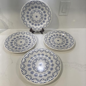 May include: A set of four white ceramic plates with a blue floral and swirl design. One plate is displayed on a wooden stand. The plates have a circular pattern of blue flowers and swirls around the edges.