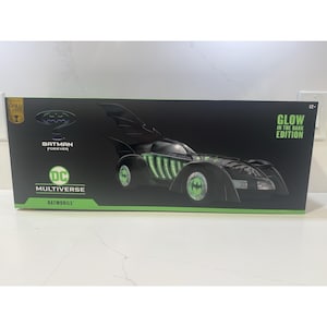 May include: Black Batmobile toy car from the Batman Forever movie, with green glowing accents. The box features the DC Multiverse logo and the words "Glow in the Dark Edition".