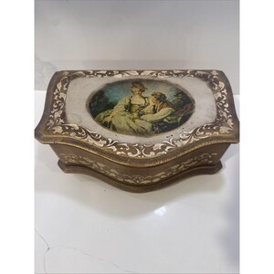 May include: A vintage jewelry box with a rectangular shape and curved edges. The box is primarily brown with gold trim and decorative floral patterns. The lid features an oval image of a couple in a garden setting. The box is likely made of metal.