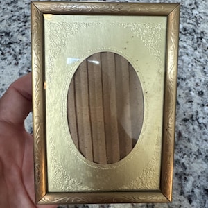 May include: A vintage gold-colored picture frame with an oval opening. The frame has an ornate, embossed design around the edges and a cardboard backing. The frame is rectangular and appears to be made of metal.