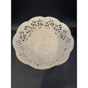 May include: A white porcelain bowl with a scalloped edge and intricate openwork design. The bowl features a raised floral pattern in the center and a gold rim. This decorative bowl is ideal for serving or display.