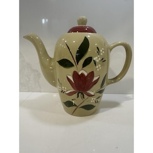May include: A beige ceramic teapot with a burgundy lid and handle. The teapot is decorated with a large burgundy flower, green leaves, and small white flowers. The spout is curved and the handle is a large loop.