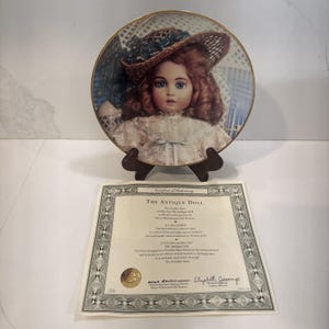 May include: A decorative plate featuring a portrait of a doll with blue eyes and red hair, wearing a hat and a white dress. The plate is displayed on a brown stand, with a certificate below. The plate has a gold rim.