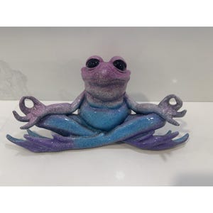 May include: A whimsical frog figurine in a seated yoga pose. The frog is painted in shades of purple and blue, with a speckled finish. Its eyes are black, and it has its arms and legs in a meditative position.