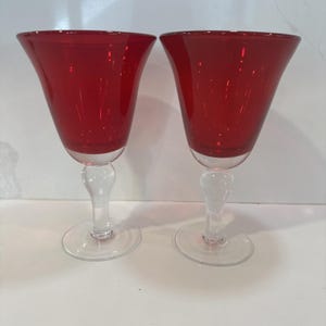 May include: Two red wine glasses with a clear, bulbous stem and a wide, flared bowl. The glasses sit on a clear, circular base. The red color of the glass is vibrant and eye-catching, perfect for a special occasion.