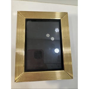 May include: A rectangular gold-colored picture frame with a black interior. The frame has a ridged design around the edges. The frame is sitting on a white surface.