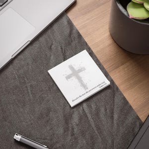Könnte beinhalten: Ein weißer quadratischer Notizblock mit einem grauen Kreuzdesign und dem Text "blessed is the man who perseveres under trial. -James 1:12". Der Notizblock liegt auf einer dunkelgrauen Oberfläche.