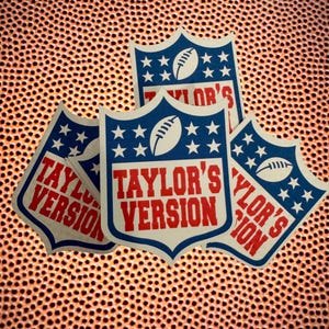 Peut inclure: Quatre autocollants en forme d'écu, avec un ballon de football et des étoiles sur fond bleu. Le texte "TAYLOR'S VERSION" est en rouge. Les autocollants sont disposés sur un fond orange texturé.