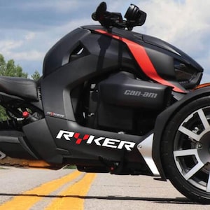 May include: A black and red Can-Am Ryker three-wheeled motorcycle. The vehicle has a black body with red accents and the word "Ryker" in white and red lettering. The motorcycle is parked on a road with yellow lines.