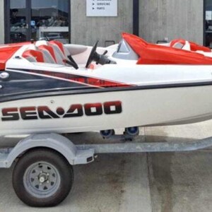 Puede incluir: Un barco Sea Doo Speedster 200 blanco y negro sobre un remolque. El barco tiene detalles y asientos rojos. El nombre del barco, "SEA DOO 200", se muestra en letras negras grandes en el lateral.